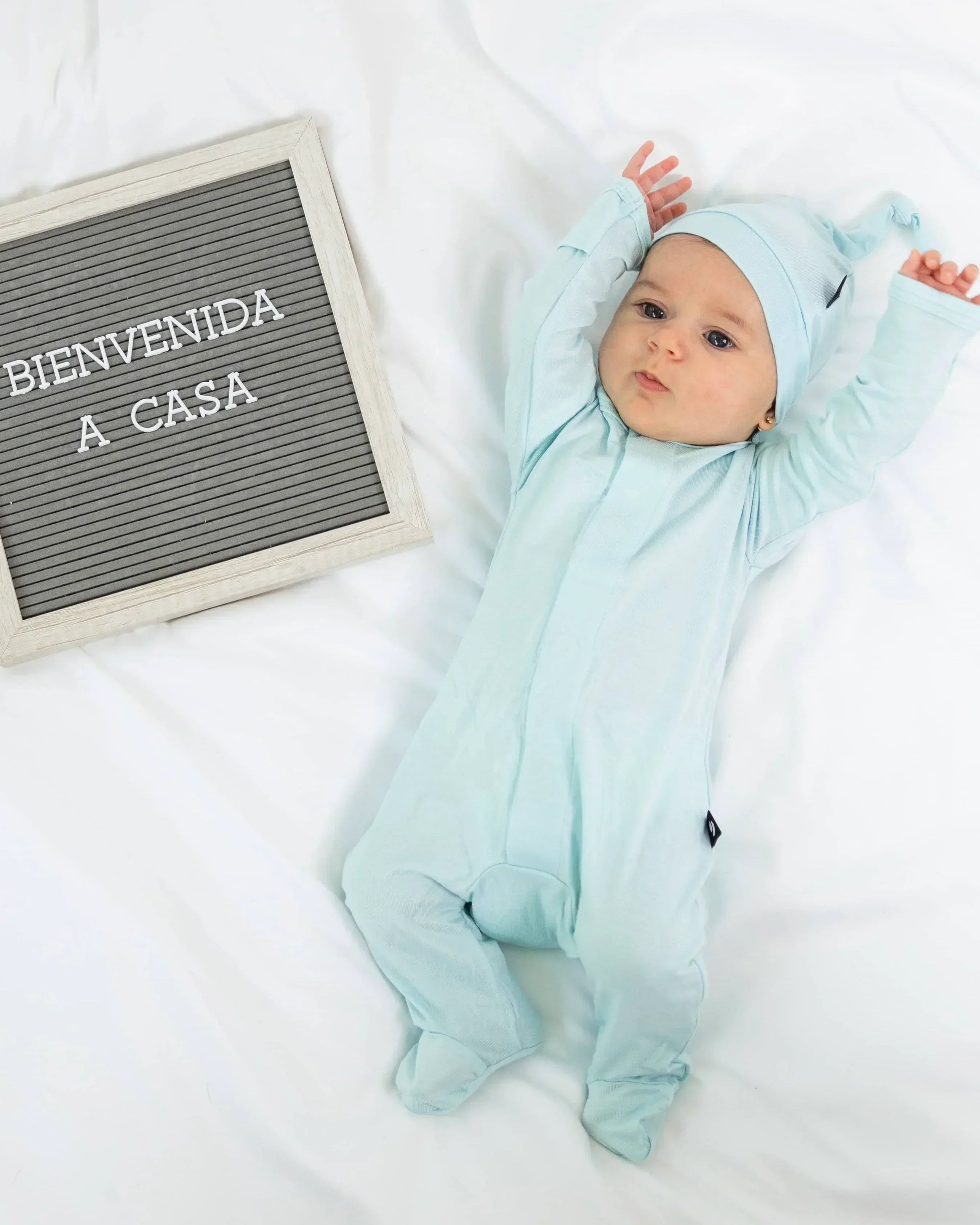 Baby in light blue onesie and hat lying on white bedding with 'Bienvenida a casa' sign