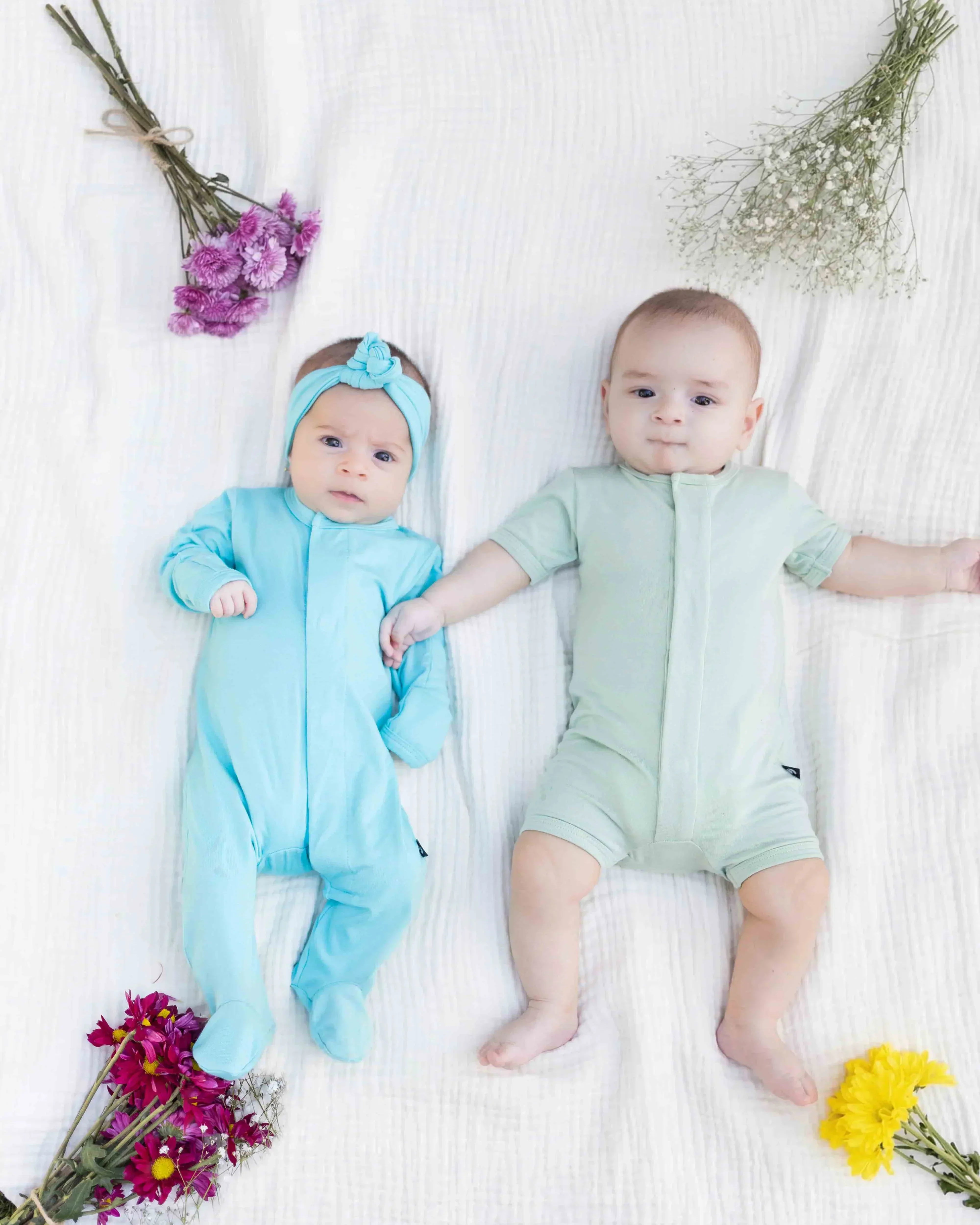 Two babies in pastel baby clothes lying on a white blanket surrounded by colorful flowers.