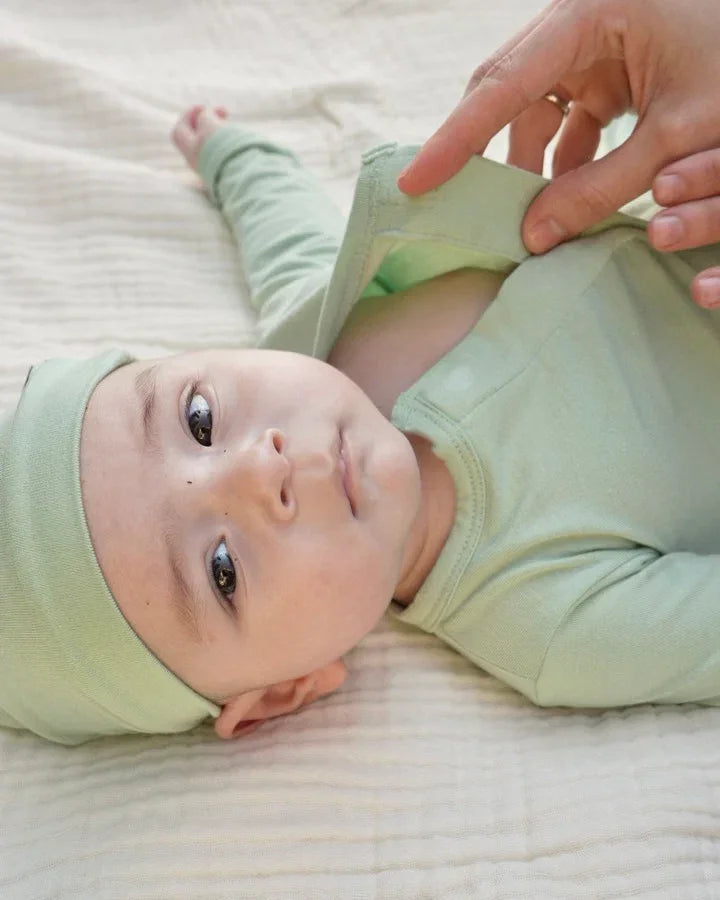 Baby in soft green onesie and hat lying on a bed, with adult fastening the outfit
